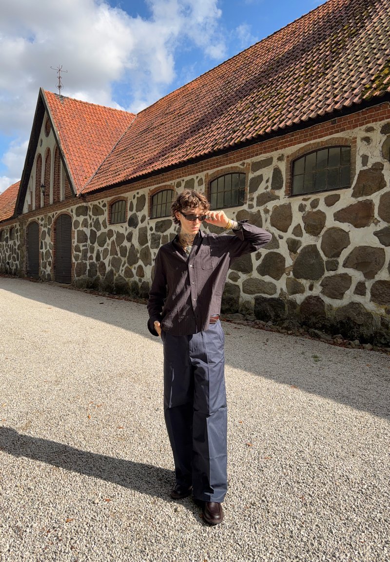Personne portant des lunettes de soleil, une chemise sombre, et un pantalon bleu large, debout sur du gravier près d'un bâtiment en pierre avec un toit en tuiles rouges sous un ciel partiellement nuageux.