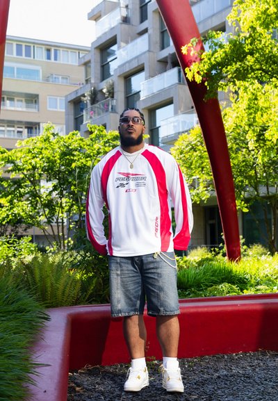 Camisa blanca de manga larga con acentos rojos, con el logo "PEGADOR", combinada con pantalones cortos de mezclilla y zapatillas blancas. Follaje verde y edificios modernos al fondo.
