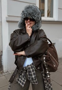 Giacca di pelle marrone con cappuccio foderato in pelliccia grigia, abbinata a una camicia di flanella a quadri legata in vita e una borsa in camoscio marrone.