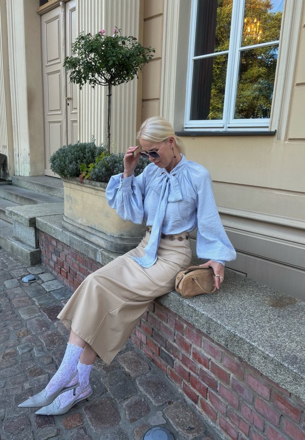 Woman with blonde hair wearing sunglasses, light blue blouse, beige skirt, and white lace socks sitting on a stone ledge outside a building.