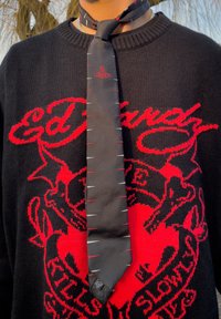 Personne portant un pull noir avec un texte graphique rouge et une cravate noire présentant de petits motifs ressemblant à des points de couture en blanc et rouge, ainsi qu'un logo rouge près du nœud.