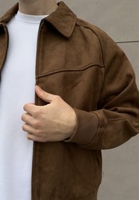 Veste en daim marron avec col, fermeture éclair et poignets élastiqués. Portée sur un t-shirt blanc, avec un design simple et une texture lisse.