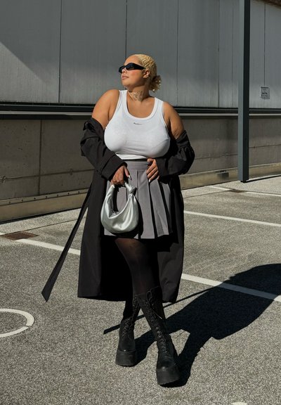 Top sin mangas blanco con logo, falda plizada gris, medias opacas negras y botas gruesas negras con cordones. Abrigo largo negro y bolso plateado.