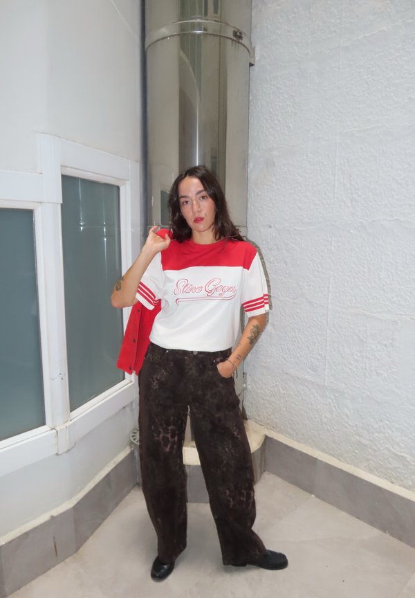 Mujer con cabello oscuro lleva camisa roja y blanca y pantalones oscuros con estampado, está de pie en el interior cerca de una tubería metálica y paredes de color claro.