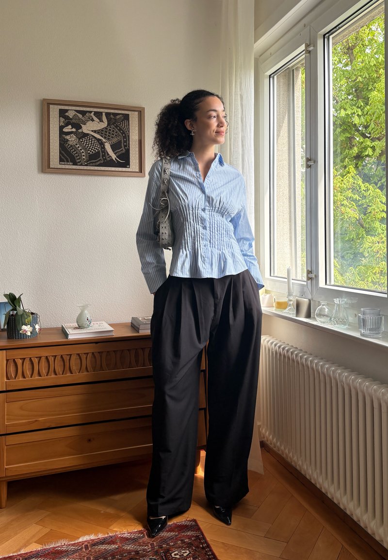Femme en blouse bleue rayée et pantalon large noir debout près de la fenêtre dans une pièce ensoleillée avec une commode en bois et une œuvre d'art encadrée sur le mur.