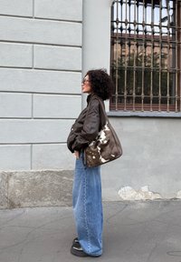 Kobieta z kręconymi włosami, ubrana w ciemną skórzaną kurtkę, szerokie dżinsy oraz buty na platformie. Niesie dużą torbę na ramię w brązowo-białe wzory.