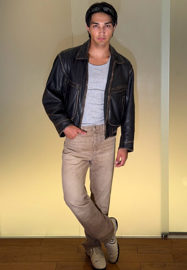 Jeune homme debout à l'intérieur portant une veste en cuir noire, une chemise grise, un pantalon beige, des baskets blanches et des lunettes de soleil sur la tête.