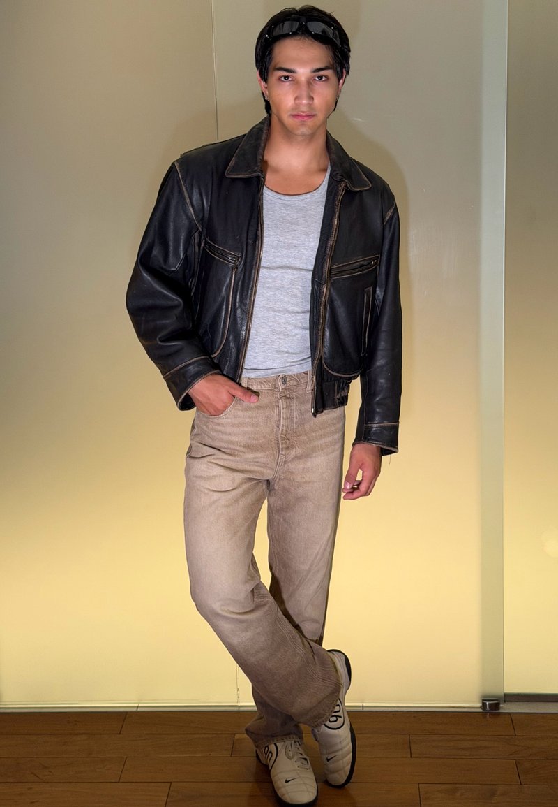 Jeune homme debout à l'intérieur portant une veste en cuir noire, une chemise grise, un pantalon beige, des baskets blanches et des lunettes de soleil sur la tête.