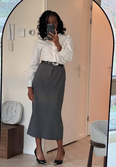 Camisa blanca de botones, falda lápiz gris, tacones negros con puntas metálicas, cinturón texturizado. De pie frente a un espejo de cuerpo entero.