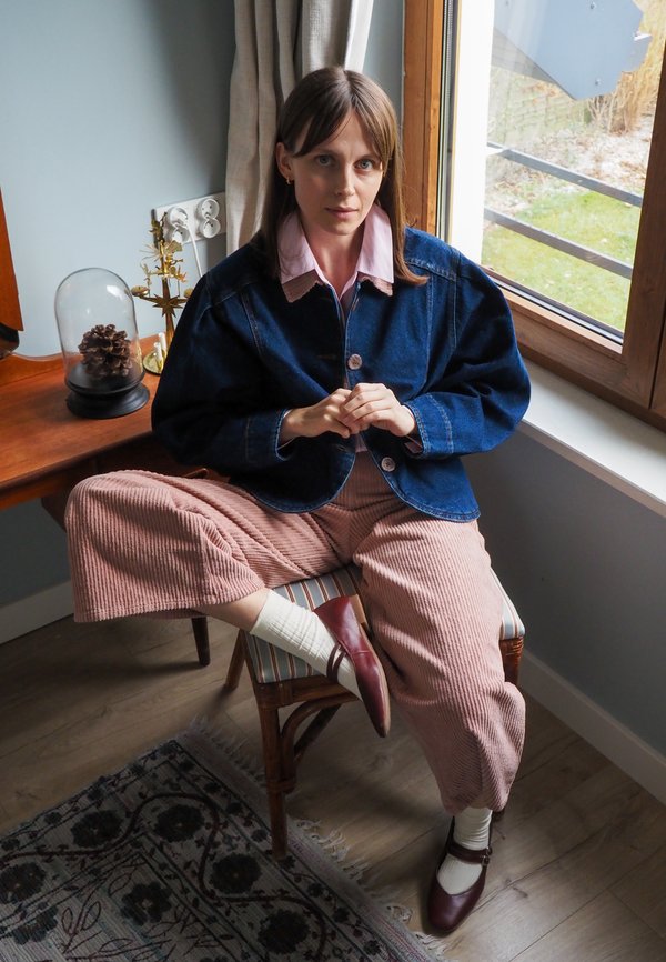 Jeansowa kurtka z bufiastymi rękawami, różowe sztruksowe spodnie w szerokim kroju, bordowe buty i białe skarpetki, siedząca przy oknie z naturalnym światłem.
