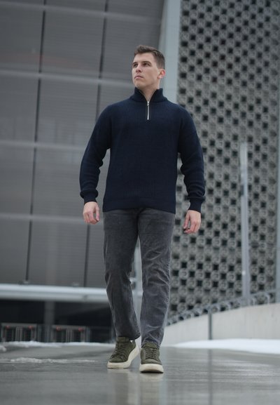 Joven caminando al aire libre con un suéter azul marino con cremallera, jeans grises y zapatillas verdes, frente a un edificio moderno.