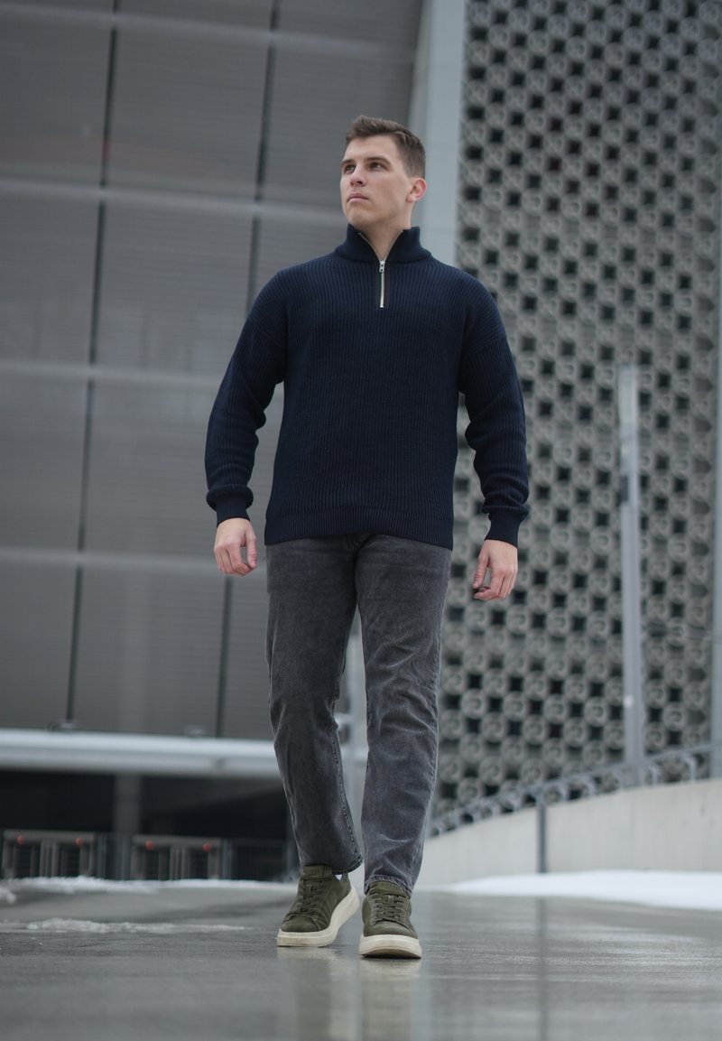 Jeune homme marchant en plein air portant un pull zippé bleu marine, un jean gris et des baskets vertes, devant un bâtiment moderne.
