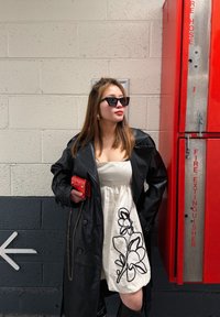 Femme en manteau de cuir noir sur une robe blanche à motifs floraux, tenant un portefeuille rouge, portant des lunettes de soleil noires, debout près d'une armoire à extincteur et d'un panneau fléché mural.