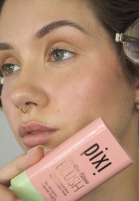Blush Pixi On-the-Glow dans un tube à deux couleurs ; section supérieure rose, base verte. Modèle le tenant près d'un visage légèrement blushé avec un look naturel.