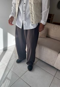 Biela dlhou rukávom košeľa pod svetlo béžovým prešívaným vestom, v kombinácii s voľnými tmavohnedými nohavicami a čiernymi papučami, stojaca v interiéri.
