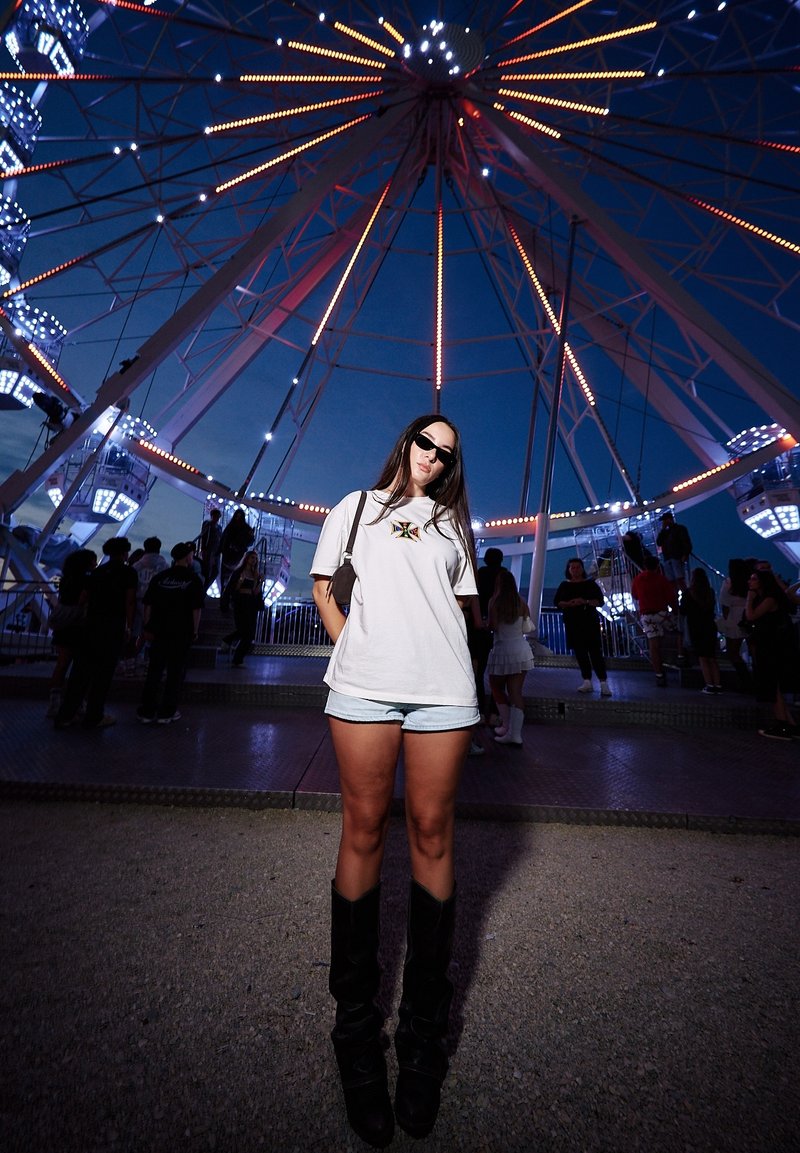 Jeune femme portant des lunettes de soleil, une chemise blanche, un short et des bottes noires se tient devant une grande roue éclairée au crépuscule, avec d’autres personnes à proximité.