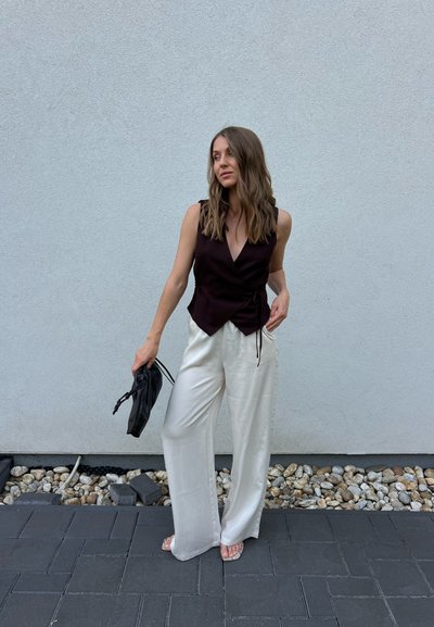 Mujer con cabello ondulado que lleva un top negro sin mangas y pantalones blancos de pierna ancha, sosteniendo un bolso negro de mano, de pie sobre suelo de piedra y baldosas frente a una pared gris.