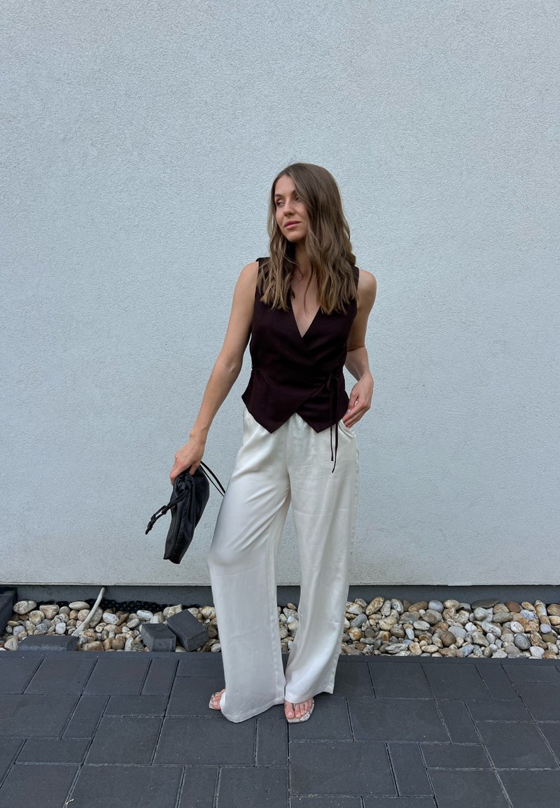 Femme aux cheveux ondulés portant un haut noir sans manches et un pantalon blanc à jambes larges, tenant une pochette noire, debout sur un sol en pierre et carreaux devant un mur gris.