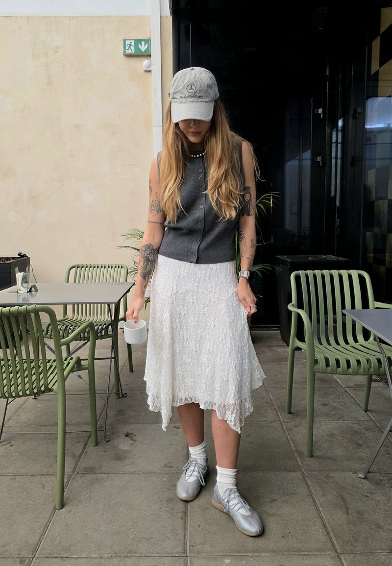 Haut sans manches gris, jupe en dentelle blanche avec ourlet asymétrique, chaussures argentées et chaussettes blanches. Tenant une tasse blanche, debout sur un sol carrelé.