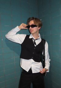 Melna veste pār baltu svītrainu krekla, melnas bikses un tumšas saulesbrilles. Pamanāmas detaļas ir rakstaina bandana un melna plecu soma.