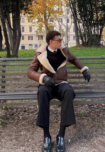 Chaqueta de borrego marrón forrada con guantes negros, pantalones negros y mocasines negros brillantes. Sentado en un banco con follaje otoñal de fondo.