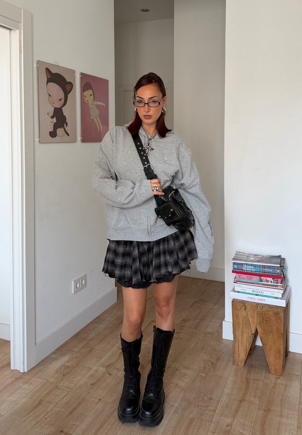 Femme avec des lunettes portant un sweat-shirt gris oversize, une jupe noire à carreaux, des bottes noires montantes et un sac bandoulière, debout dans une pièce au sol en bois.