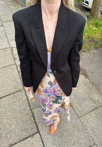 Blazer negro ajustado sobre un vestido midi floral en crema con patrones morados y naranjas. Tacones nude de punta naranja y un bolso ligero.