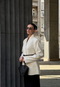 Witte blazer met brede mouwen, ceintuur in de taille, gecombineerd met een zwarte handtas. Model draagt donkere zonnebril, met een soepele textuur en een taillering die goed past.