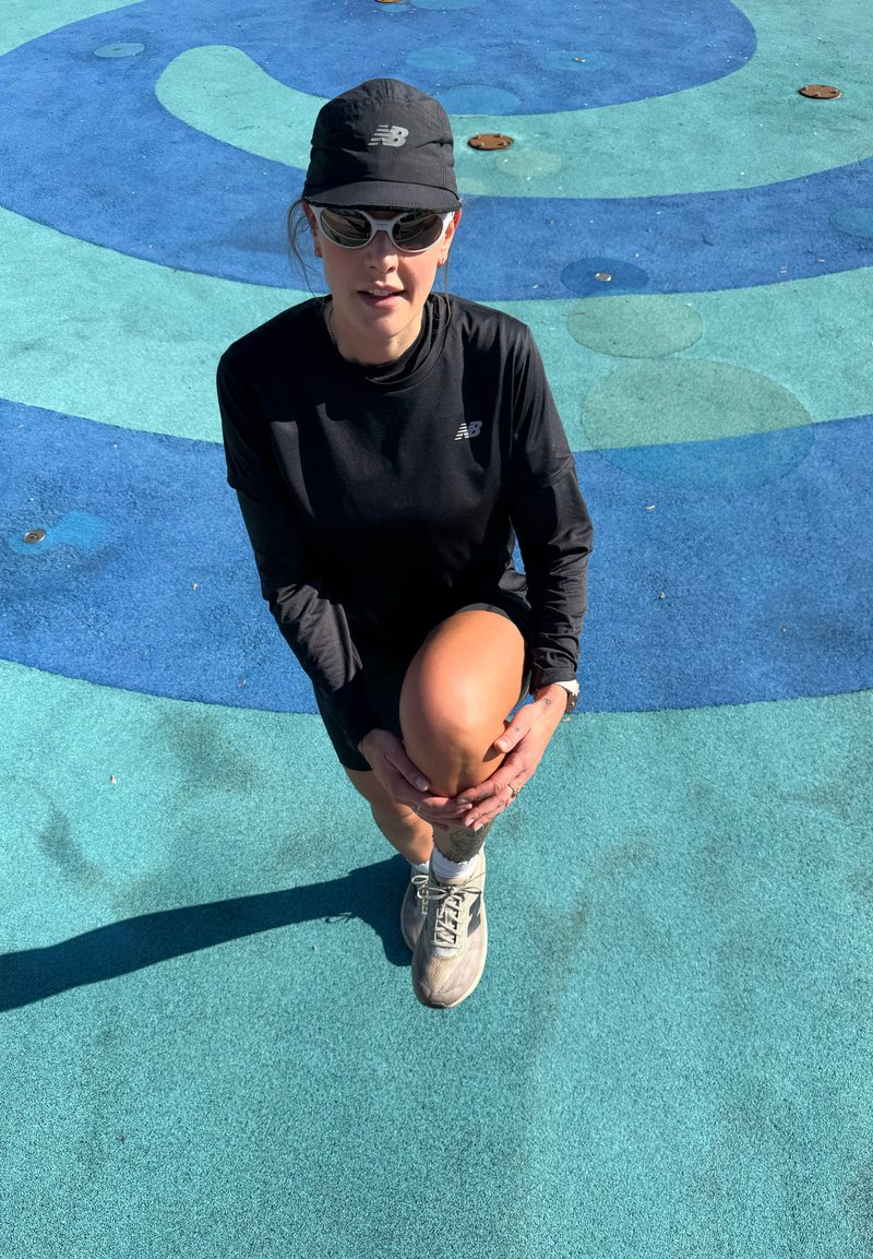 Žena v černém sportovním oblečení a kšiltovce natahuje levou nohu na modrém vzorovaném povrchu na slunci.