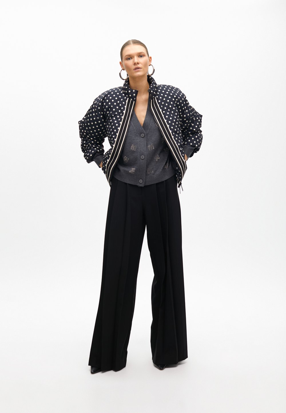 Giacca bomber imbottita con pois bianchi e neri, cardigan grigio e pantaloni larghi neri. Orecchini a cerchio grandi aggiungono un tocco di dettaglio.