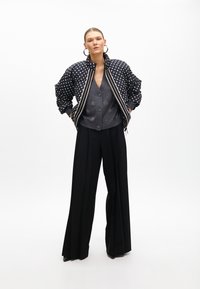 Giacca bomber imbottita con pois bianchi e neri, cardigan grigio e pantaloni larghi neri. Orecchini a cerchio grandi aggiungono un tocco di dettaglio.