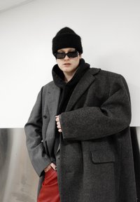 Personne portant un manteau gris trop grand par-dessus un sweat à capuche noir, un bonnet noir, des lunettes de soleil rectangulaires et un pantalon rouge, debout contre un mur blanc.