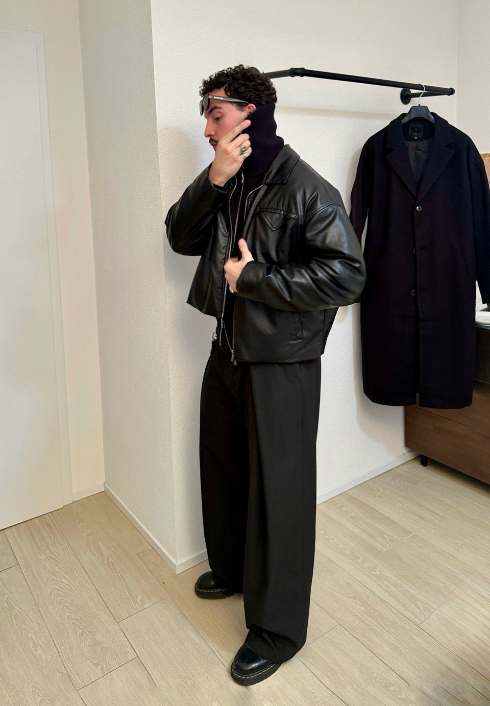 Un homme portant une veste en cuir noire, un pantalon large noir et des lunettes de soleil ajuste son col roulé dans une pièce minimaliste avec un manteau noir accroché à un porte-manteau.