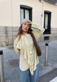 Κρεμ καφέ, ριχ�τό πουκάμισο με γιακά, συνδυασμένο με ανοιχτό μπλε παντελόνι με φαρδιά μπατζάκια. Αξεσουάρ περιλαμβάνουν ένα πράσινο σκούφο και έναν καφέ ώμου.