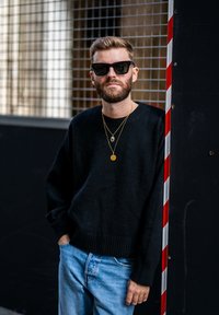 Suéter negro de punto con un corte holgado, combinado con jeans de color azul claro. Dos collares de oro adornan el cuello. El fondo presenta un poste a rayas.