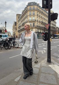 Uformell antrekk med grå hettejakke med glidelås, hvit singlet, forkle med blonder, vide grå bukser og mønstret veske. Urban gateomgivelse.