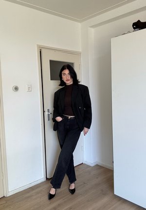 Mujer con cabello negro está de pie en una habitación vistiendo un blazer negro, top oscuro, pantalones negros y tacones negros, apoyada contra una puerta cerrada.