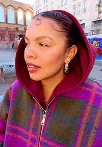 Felpa con cappuccio in peluche multicolore con motivi a quadri nei toni del marrone rossiccio, verde e viola. Cerniera argentata e grandi orecchini decorati.