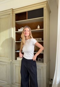 T-shirt blanc à manches courtes associé à un pantalon large plissé de couleur sombre. En arrière-plan, des meubles en bois clair avec des étagères et des objets décoratifs.