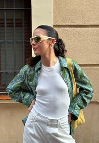 Camisa verde com padrão paisley sobre uma blusa branca ajustada, combinada com calças brancas de cintura alta e uma bolsa amarela; usando óculos de sol de cor clara.
