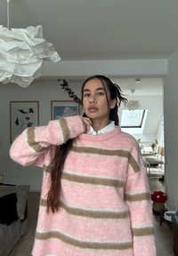 Pull rose duveteux avec des rayures horizontales blanches et marron, col rond et manches longues. Superposé sur une chemise blanche à col.