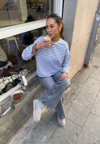 Chemise rayée bleu et blanc à manches longues, jean gris évasé, bottines beiges. Assise, tenant une boisson, avec une assiette de gâteau à côté.