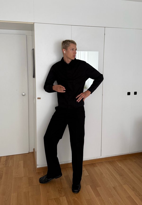 Joven vestido de negro con auriculares puestos, de pie con una mano en la cadera en una habitación con paredes blancas y suelo de madera.