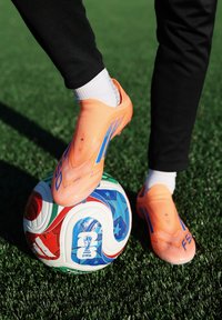 Personne portant des crampons orange contrôle un ballon de football de la Coupe du Monde de la FIFA sur un terrain synthétique vert.