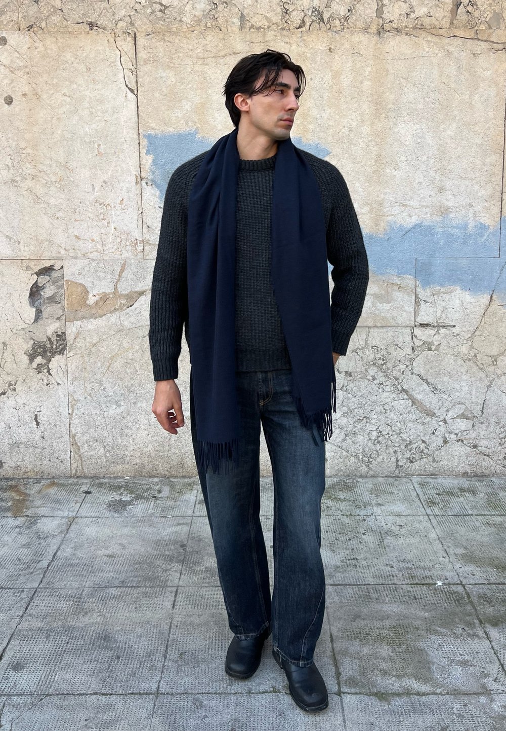 Homme portant un pull en maille foncé, une écharpe marine, un jean délavé et des chaussures noires, debout sur un trottoir carrelé devant un mur en pierre texturé.