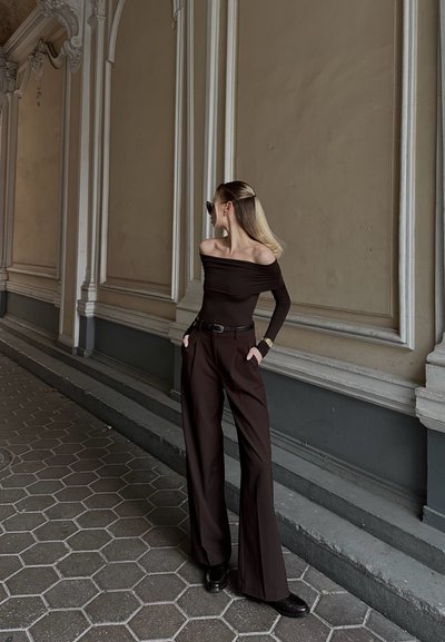 Mujer con un top marrón oscuro de hombros al descubierto y pantalones de pierna ancha, de pie con las manos en los bolsillos, mirando hacia la izquierda, frente a una pared beige ornamentada.