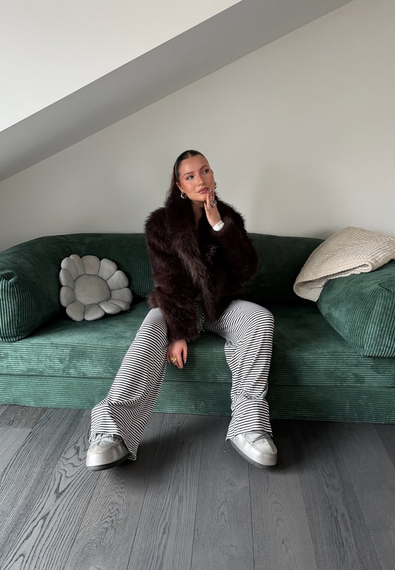 Femeie cu haină de blană închisă la culoare și pantaloni cu dungi, stând pe o canapea verde cu pernă în formă de floare și pătură pliată într-o cameră modernă.