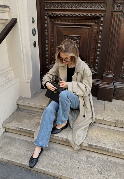 Gabardina beige claro sobre una blusa negra, sentada en unos escalones de piedra. Jeans de mezclilla y zapatos planos negros. Bolso negro con detalles en dorado.