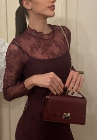 Γυναίκα με σκούρο μπορντό δαντελένιο φόρεμα που κρατά τσάντα σε αντίστοιχο χρώμα με χρυσή αλυσίδα, σκούρα περιποιημένα νύχια και μαλλιά δεμένα πίσω σε αλογοουρά.