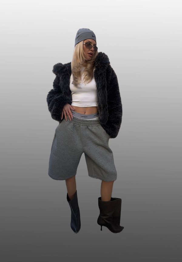 Kvinde iført grå hue, solbriller, sort pelsjakke i imiteret kaninpels, hvid crop top, grå oversized shorts og ujævne sorte ankelstøvler står mod en grå baggrund.